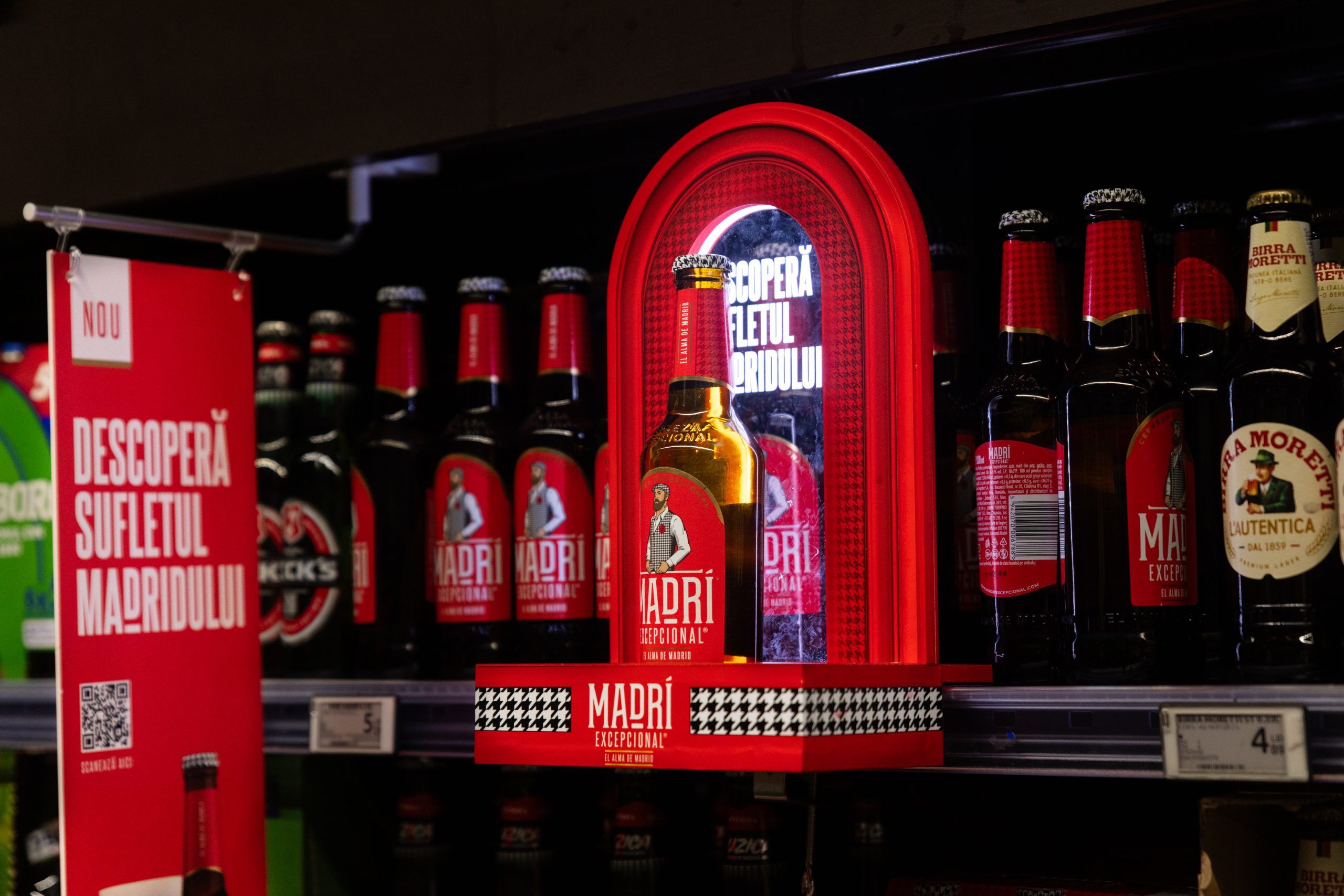 Sticlă de bere Madri Excepcional iluminată într-un glorifier roșu, expusă pe raft într-un supermarket, însoțită de afiș cu textul „Descoperă sufletul Madridului”.