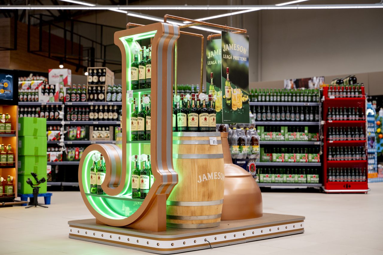 Stand promoțional Jameson într-un supermarket, cu sticle expuse într-o instalație iluminată în formă de literă J, alături de un butoi din lemn și elemente decorative de tip alambic.